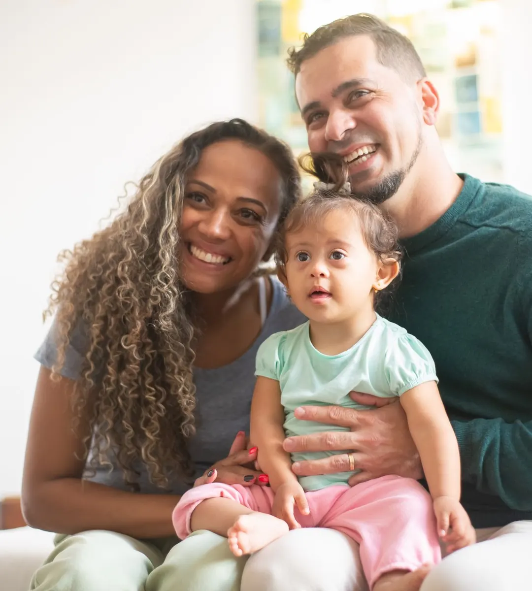 Mãe e pai sorrindo no sofá com a filha pequena no colo