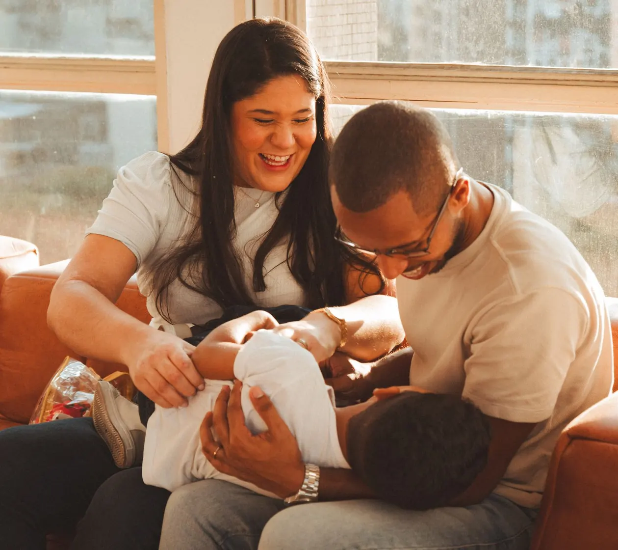 Mãe e pai sorrindo no sofá com o filho no colo, numa sala iluminada pelo sol